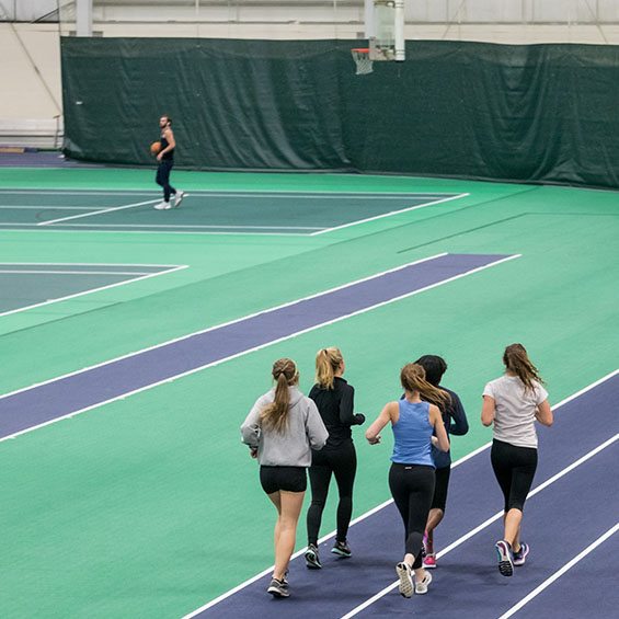 Five students jog around the Costello Sports Complex indoor track while another student can be seen in the background playing basketball.