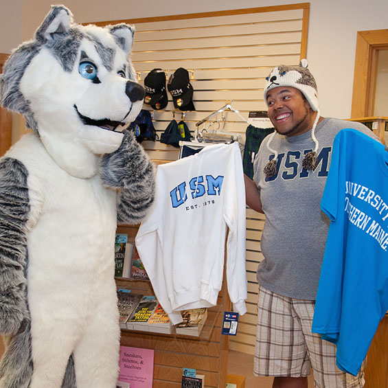 Our mascot, Champ, and a student try to decide between a USM sweatshirt and University of Southern Maine t-shirt. The student is wearing a winter hat with ear flaps that features a husky head.