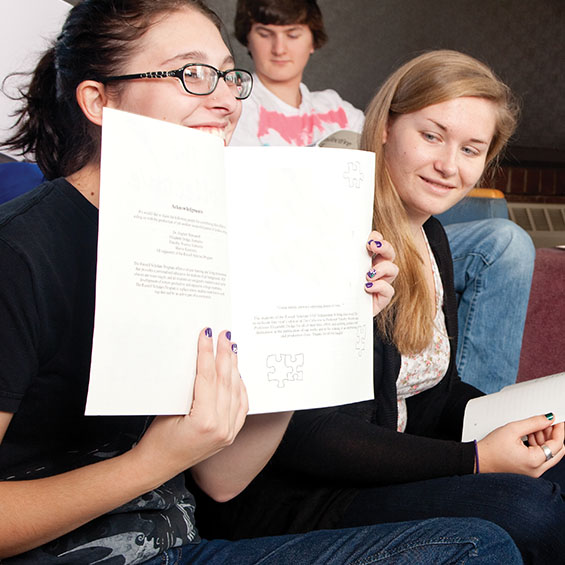 A student seated on the floor of a residence hall common space holds up an open workbook as other students in the group look on.