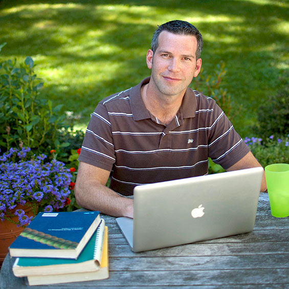 A person sits at a picnic table in their back yard. A laptop is open in front of them, between a stack of textbooks and a bright green cup.