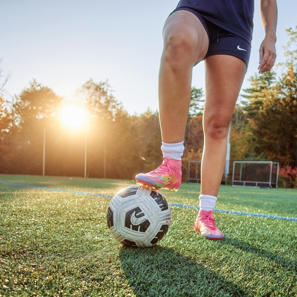 A soccer player, shown from the waist down and wearing pink cleats, rests one foot on a soccer ball.
