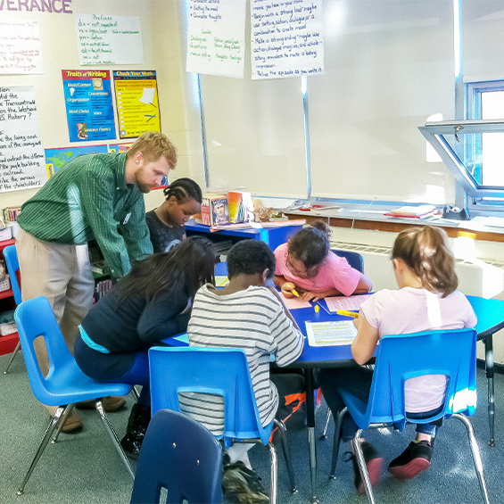 Student teacher leans over table with 5 seated elementary school students.