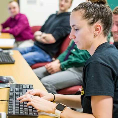 A student typing on a keyboard in a classroom while other students look on