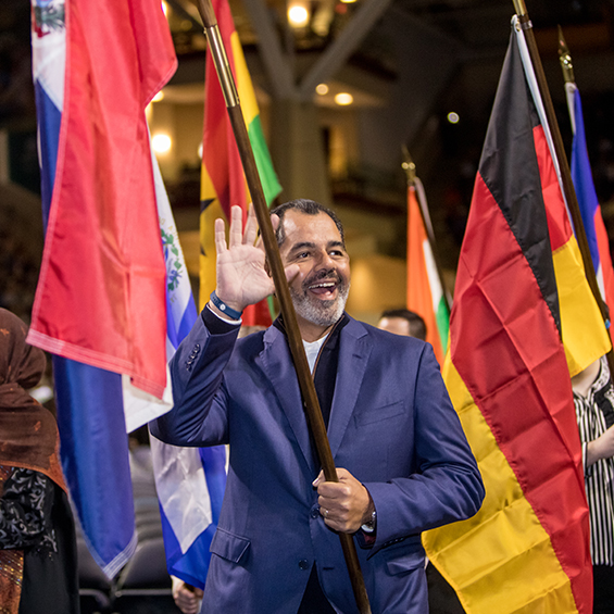 A man smiles and waves while holding a large flag, among many other large flags of various countries.