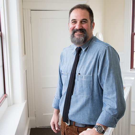 A teacher stands in a hallway, leaning against a railing, and smiles.