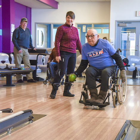 An elder in a wheelchair bowls a ball at a bowling lane next to a coach.