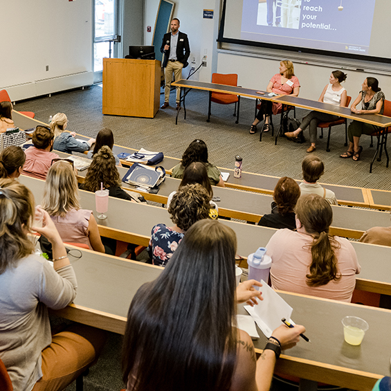 Graduate students attend their orientation in a lecture hall, with a staff member speaking at a podium.