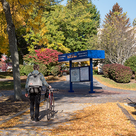 A student walks their bike on a sidewalk toward a campus map.