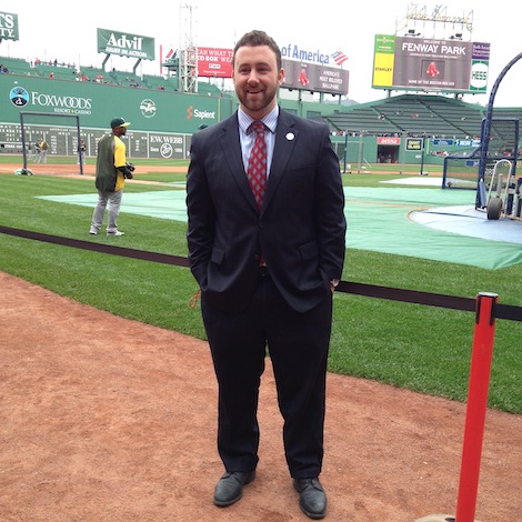 A student stands at the edges of a baseball field while wearing a suit.