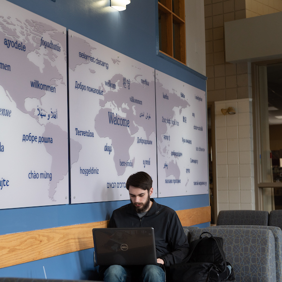 A student sits in front of a world map hanging on a wall and works on their laptop.
