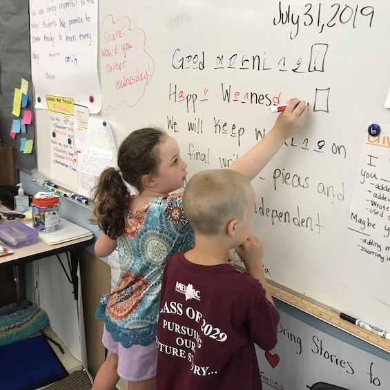 Two elementary school students write answers on a whiteboard in a classroom.