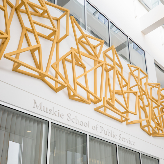 The exterior of the Muskie School of Public Service. We see the school's name written on the side, along with a geometric metal sculpture.