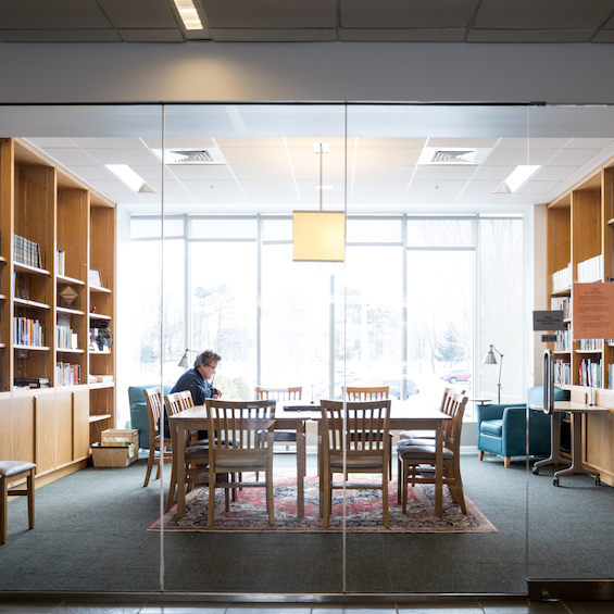 A student studies in a brightly lit private room. Glass doors, glass windows, and bookshelves surround the student as they sit at a large table.