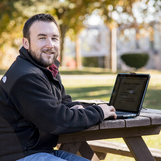 A student sits at wooden picnic table outside and studies on his laptop.