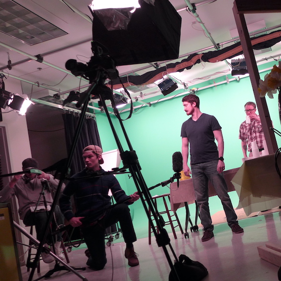 Students using camera and sound equipment film student actors filming in front of a green screen.