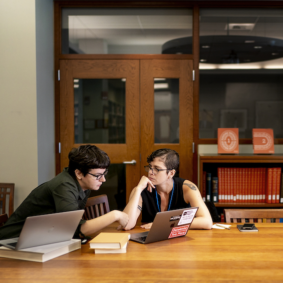 Two students study together in the library, with books and laptops on a large table.