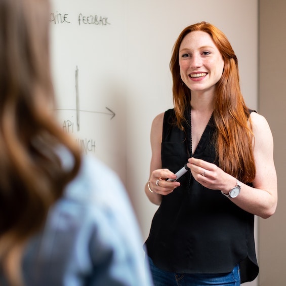 A woman with long hair and a black shirt holds a black dry erase marker and talks with a student while they both stand in front of a whiteboard.