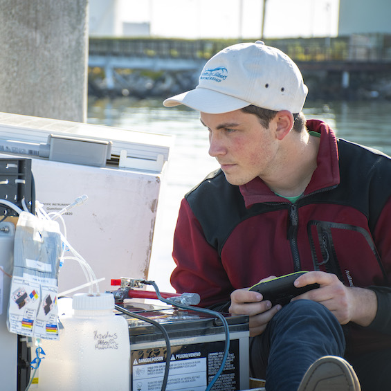 A student utilizes research equipment, sitting on a dock on the coast.