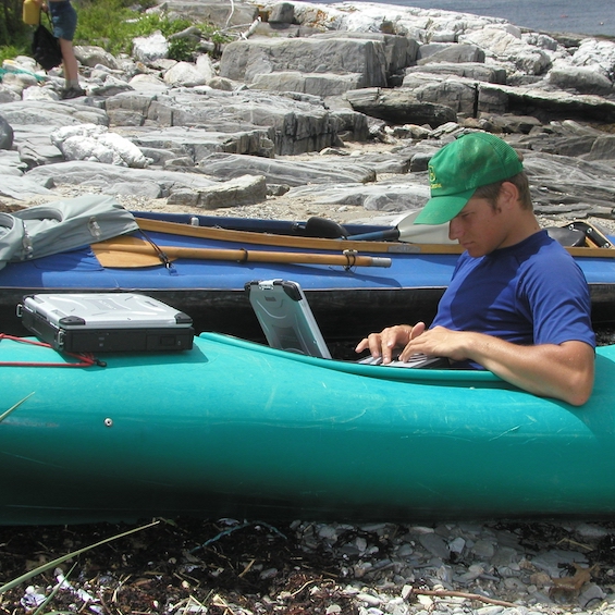 A student sits in a kayak and types on a laptop.