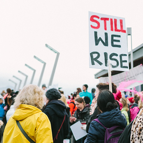 People gather at a protest outside and hold a sign reading still we rise.