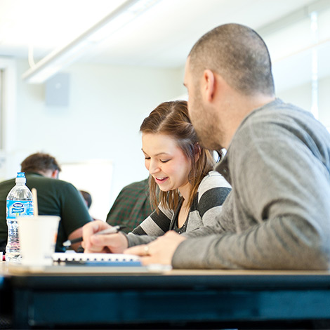 Two students have a discussion at a table in a classroom.