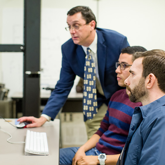 A faculty member demonstrates on a computer while two seated students watch.