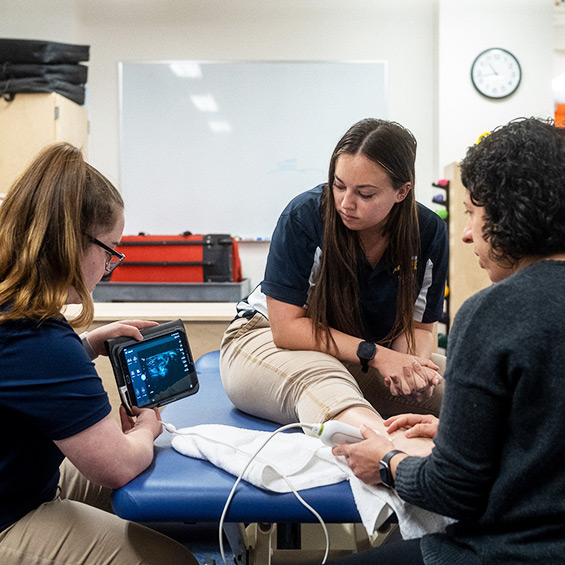A faculty member instructs students on the use of an ultrasound while one student practices using it on another's student's lower leg.