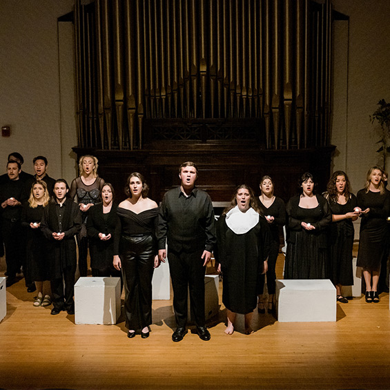 A cast of the musical Nine stand in black costumes on the stage of Corthell Hall.