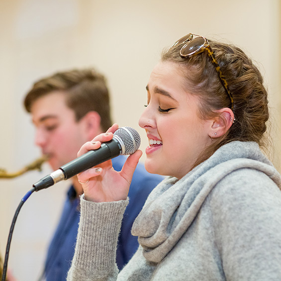 A singer sings into a microphone with her eyes closed, and in the background a student plays the saxophone.
