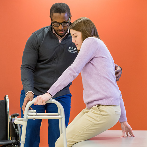 A student helps someone stand with the use of a walker.