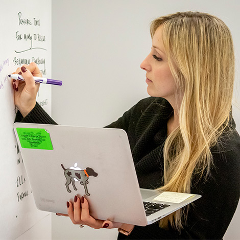 A student holds a laptop and writes on a large piece of paper that is taped to the wall.