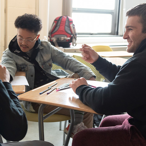 Students sit at desks in a group and talk during a class.