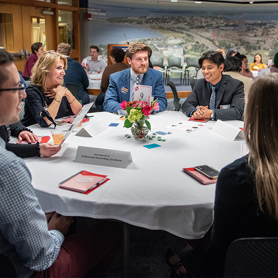 A group of students and business leaders site at a round table with notebooks, talking and smiling.