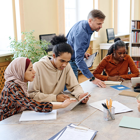 A group of adult students sit at a table, reviewing papers, while a teacher provides instruction.