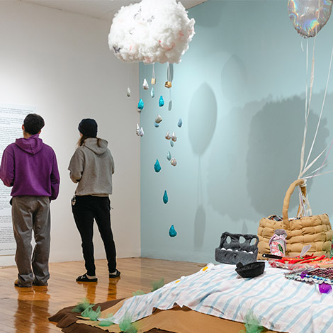 An art exhibition includes a picnic setting made of fabric, with a fabric rain cloud and rain drops hanging from the ceiling, and two people standing in the background.
