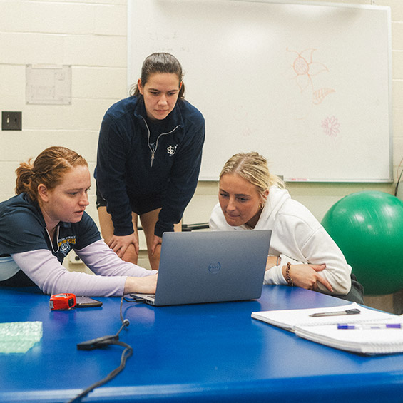 Two students sit and work at a laptop while a faculty member provides instruction, in a room with exercise equipment including a yoga ball.