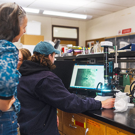 A faculty member stands behind a student who sits at a counter with computer and a microscope.