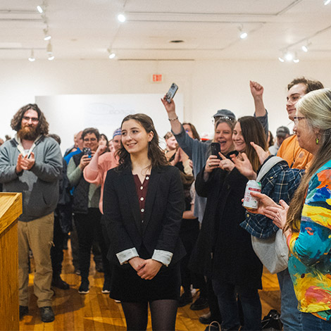 A student smiles while accepting an award, with a cheering crowd behind her.