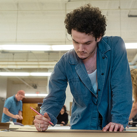 A student leans over a table with a pencil, marking cardboard.