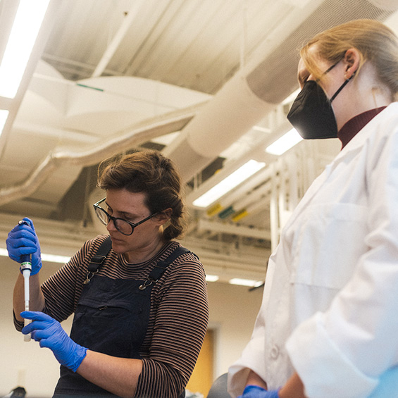 Two women work in a lab; one is using equipment while the other stands in a white lab coat and observes.