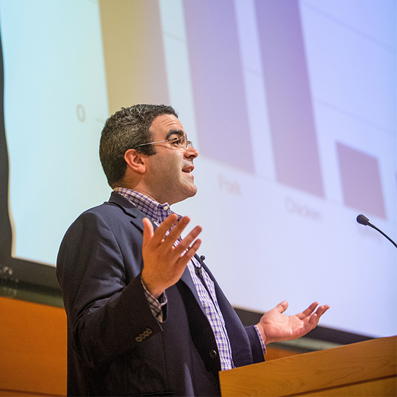 A man stands at a podium and speaks into a microphone, with a large bar chart projected on a screen behind him.