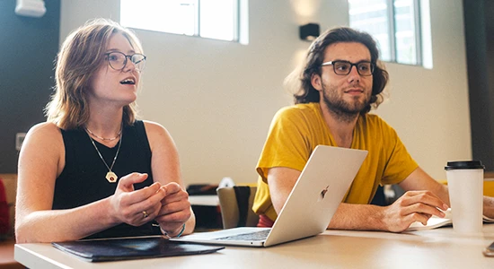 Two students sit at a table with a laptop, one student is speaking to others seated at the table outside of the picture while the other student listens.