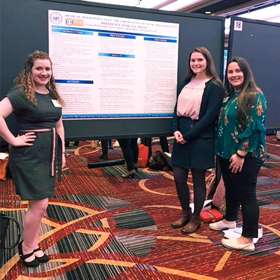 A group of three students stand in front of a large poster that includes text for an academic presentation.