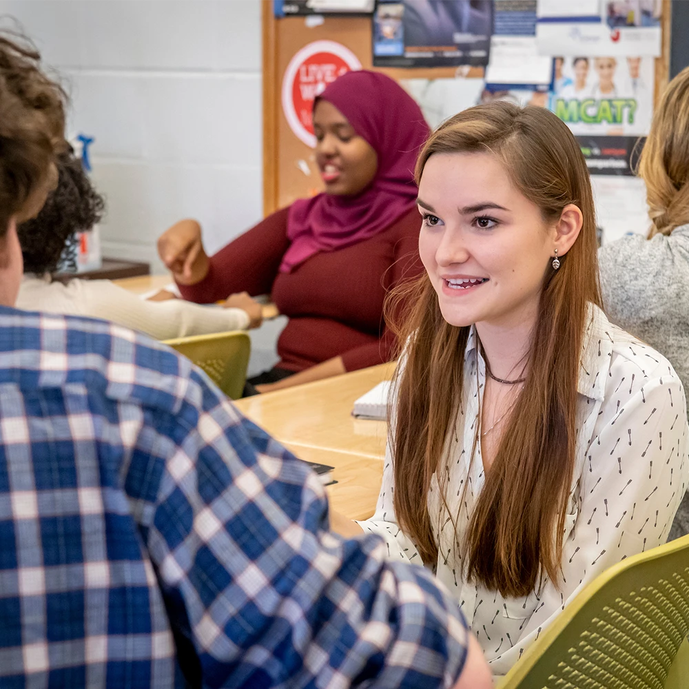 A student sits in class and smiles at another student she is talking to, with other students sitting in the background.