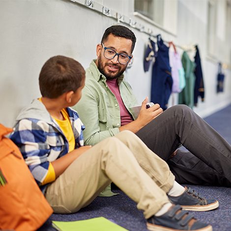 A teacher sits in a school hallway and talks to an elementary school student. Backpacks and coats hang on hooks in the background.