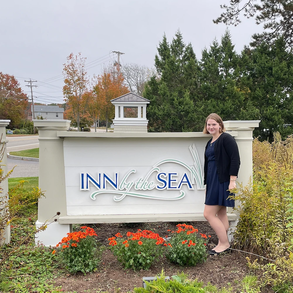 A student poses outside by a large wooden sign inscribed with "Inn by the Sea."