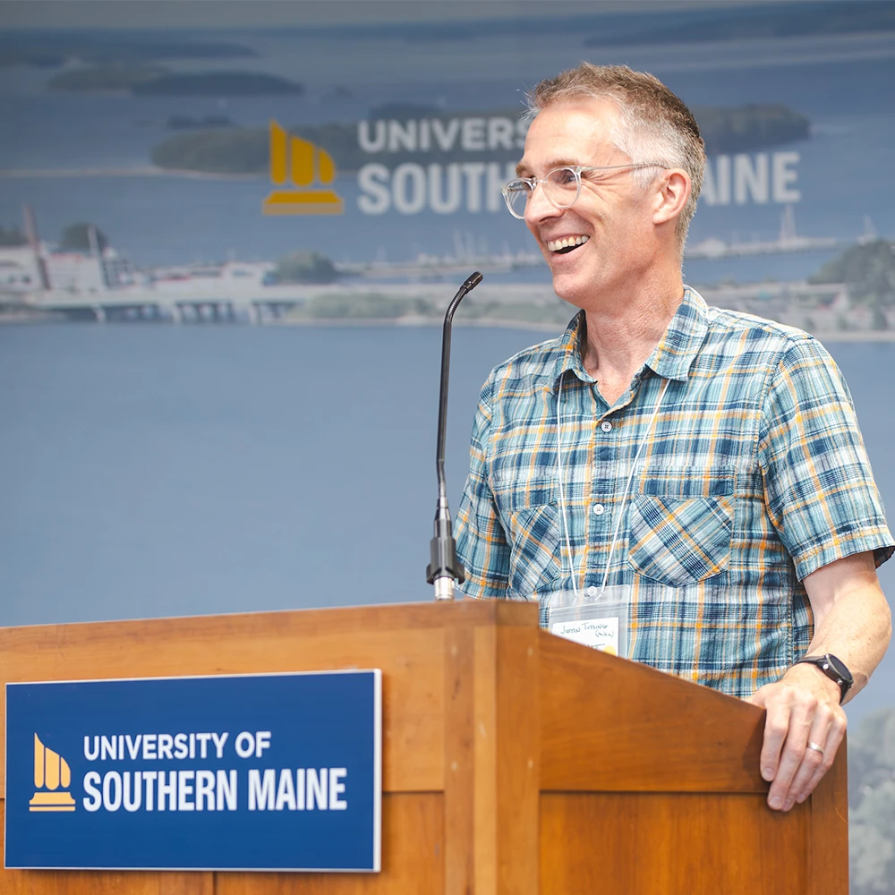 A faculty member stands at a podium and speaks into a microphone, smiling.