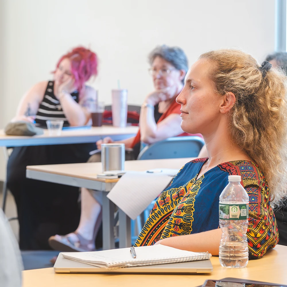 A group of three students sit at different tables and look in the same direction, listening to someone.