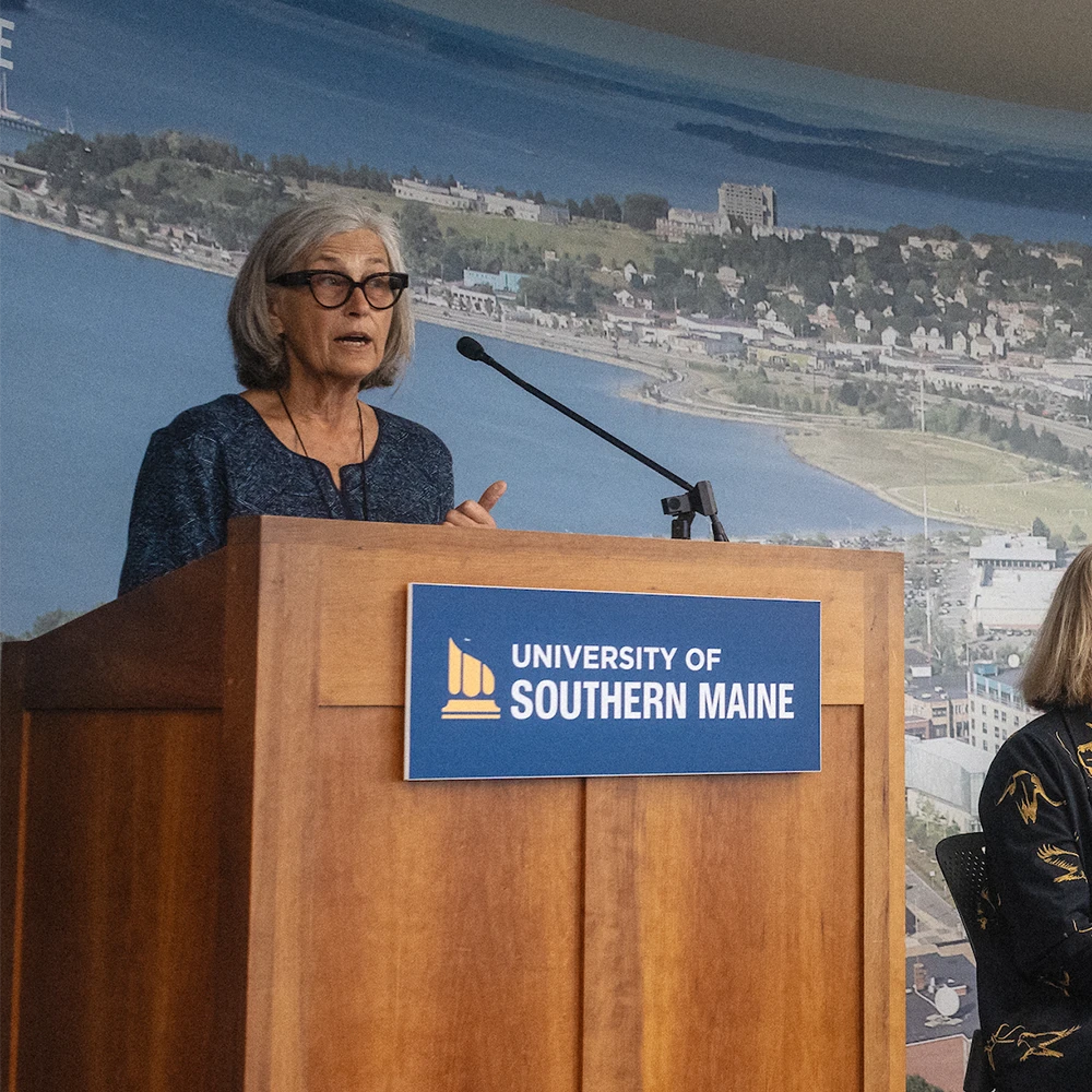 A faculty member stands at a podium and speaks into a microphone. A sign on the podium reads "University of Southern Maine."