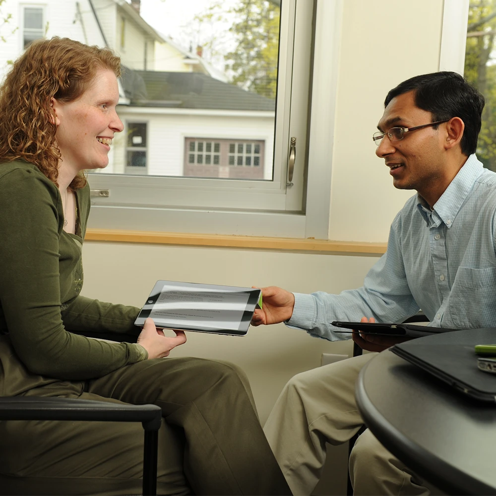 A man and woman are seated in an office and the man is handing a tablet to the woman.